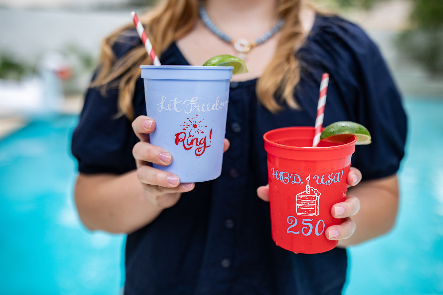 Person holding a blue and a red cup with text and straws by a pool.
