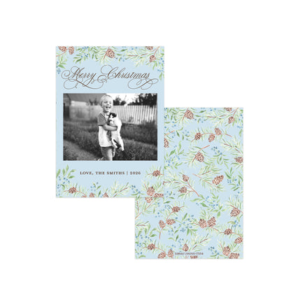 A personalized Christmas photo card and its matching envelope set. The light blue card features a floral pinecone border, a central black-and-white photo of a child holding a dog, and elegant cursive text reading 'Merry Christmas' and 'LOVE, THE SMITHS | 2026'.