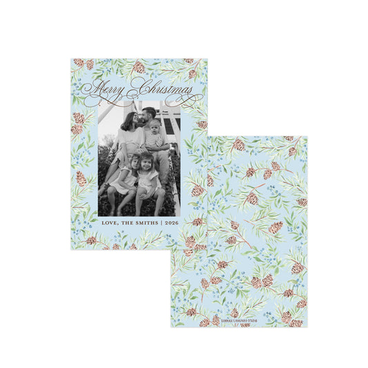 Two personalized Christmas photo cards with a light blue background, decorated with a watercolor-style floral and pinecone border. One card shows a black-and-white family photo with 'Merry Christmas' in silver script at the top, and the other shows the full repeating pattern.