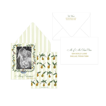 A coordinated holiday stationery set on a white background, including a striped envelope, a printed invitation card with a black-and-white couple photo framed by greenery, a matching card with pears on a striped background, and a white address label card with gold script text.