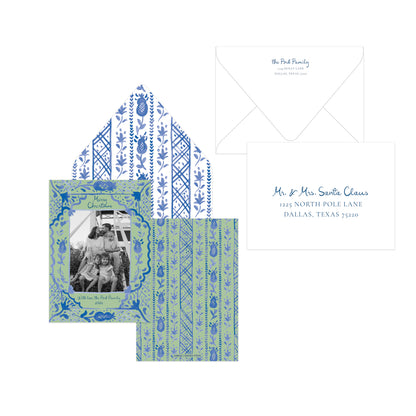 A coordinated stationery set on a white background: a square invitation card with a family photo and blue botanical border, a matching patterned envelope liner, an open white envelope, and a white address card with blue script text.